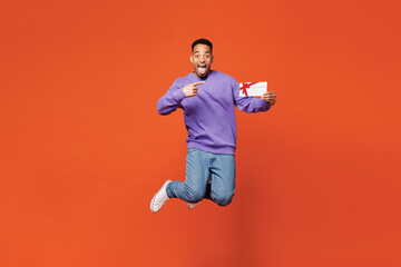 Full body young man of African American ethnicity wear purple sweatshirt casual clothes jump high hold point finger on gift certificate voucher card for store isolated on plain red orange background.