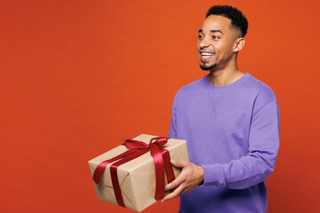 Young happy man of African American ethnicity wears purple sweatshirt casual clothes hold give present box with gift ribbon bow look aside isolated on plain red orange background. Lifestyle concept.