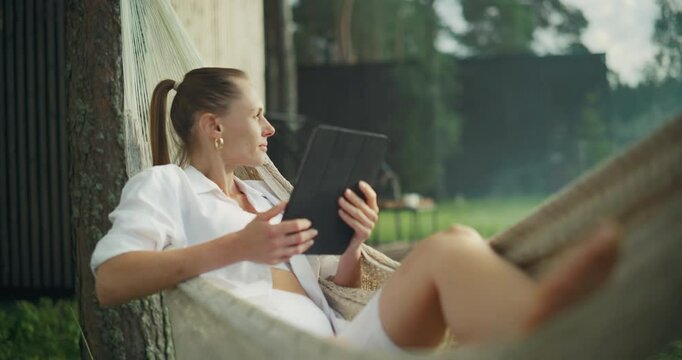 Female Enjoying Time Off From the Office, Using a Tablet Computer while Lying on a Hammock Outdoors on a Terrace of Her Country House. Young Woman Browsing Online, Shopping, Checking Social Media