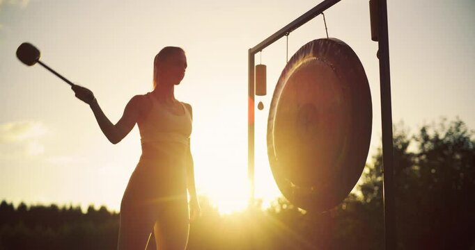 Standing in the Warm Evening Light, Serene Woman Hits a Metal Gong, Letting Its Soothing Resonance Deepen Her Relaxation. Vibrations Blend with Peaceful Outdoors, Creating a Meditative Atmosphere