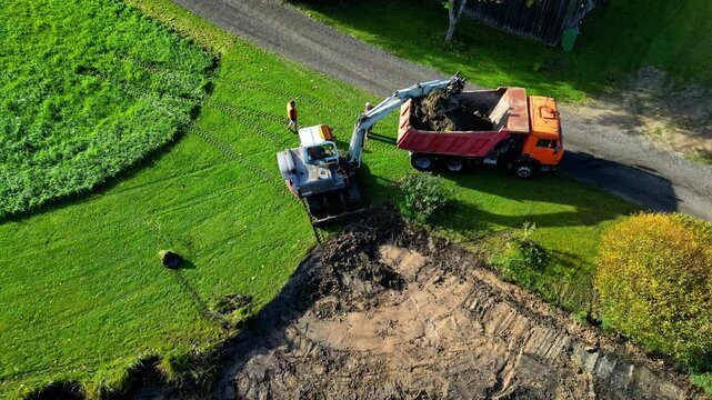 Aerial overview of excavator removing dirt from grassy lawn to prepare a new build plot, drone top down slow motion