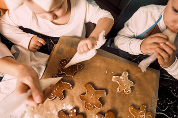 A top view of a black table with gingerbread cookies as a mother and her children decorate them with icing together.