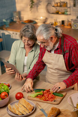 in bright kitchen an elderly woman in a light green shirt with her husband are preparing a healthy dinner the husband is cutting vegetables the wife is watching a recipe on a tablet