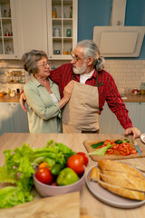 in bright kitchen an elderly woman in a light green shirt with her husband in an apron are standing in the kitchen hugging, smiling, looking at the camera