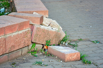 Damaged brick fence on the street, broken bricks with debris scattered on the ground, urban structure damage. Masonry wall with chipped bricks and concrete,  hazardous environment in public space
