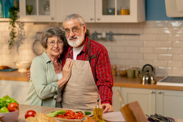 in bright kitchen an elderly woman in a light green shirt with her husband in an apron are standing in the kitchen hugging, smiling, looking at the camera