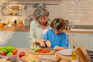 in a bright kitchen an elderly woman in a light green shirt teaches her grandson in blue T-shirt to cut vegetables for a salad