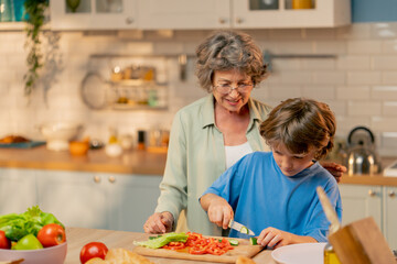 in a bright kitchen an elderly woman in a light green shirt teaches her grandson in blue T-shirt to cut vegetables for a salad