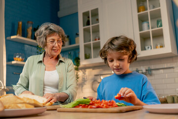 close up in a bright kitchen an elderly woman in a light green shirt teaches her grandson in blue T-shirt to cut vegetables for a salad