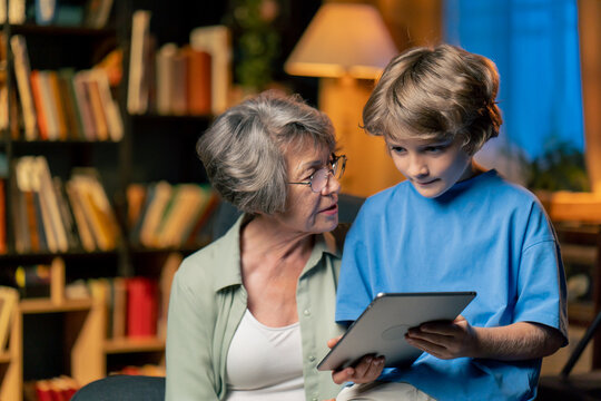 a warm light of living room with library big blue armchair elderly woman sitting with grandson watching news from tablet modern grandmother - Powered by Adobe
