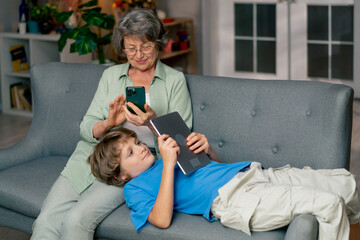 Obraz premium bright living room with sofa grandmother sitting on sofa grandson lying and watching cartoons on tablet happy family