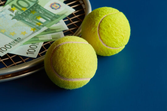 Two yellow tennis balls next to a racket with hundred euro bills on it, blue background, close-up, winnings, prize money in tennis, sports betting.