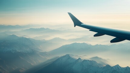 Fototapeta premium Airplane wing is flying over a mountain range covered with snow. The sun is rising, creating a warm, hazy glow over the landscape