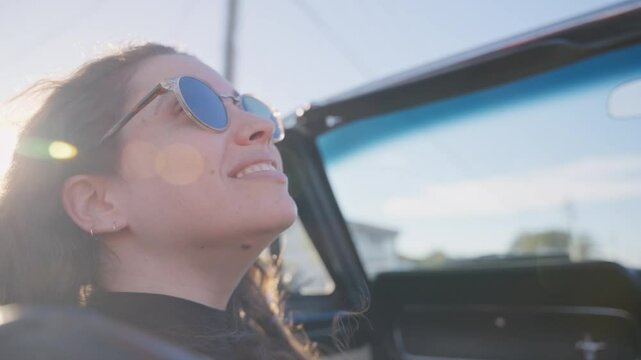 Woman smiling and enjoying ride in a vintage convertible car on sunny road trip. Young female sitting with happy face in front seat in right-hand side steering car in Australia residential city street