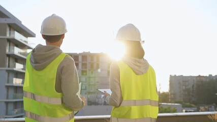 Two engineers are standing on a construction site wearing hard hats and safety vests looking at a building