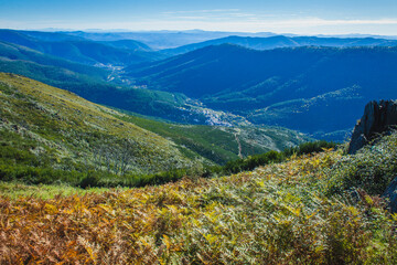 a panoramic view of a lush, expansive valley surrounded by layered mountains under a bright blue sky, with ferns and greenery in the foreground