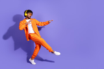 Full length photo of handsome good mood guy wear orange suit listen music earphones dancing emtpy space isolated violet color background