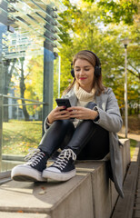 beautiful girl in wireless headphones with a phone on the street in autumn, using smartphone and headphones, chatting, listen  to music