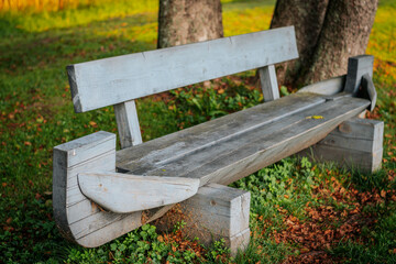 A rustic wooden bench made of rough-hewn wood sits in a grassy park area shaded by trees. The bench has unique boat-like ends that enhance its charm in its natural setting.