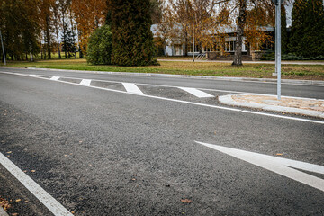 Obraz premium A quiet, empty asphalt road with a white directional arrow and painted lane markings. The scene shows fall foliage and trees in the background, with a nearby building partially visible.