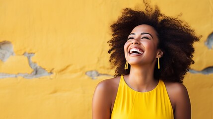 A young Black woman with curly hair laughs against a yellow wall.