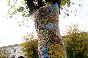A tree wrapped in colorful crochet yarn art, showcasing vibrant patterns against a backdrop of an urban park. A unique blend of nature and creative street art in autumn