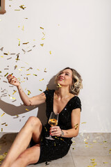 Beautiful happy woman in glamorous sparkling outfit with champagne glass celebrating, sitting at festive birthday or New Year's party surrounded by colorful confetti against white background