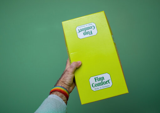 Paris, France - Feb 24, 2024: A hand holding a bright yellow Finn Comfort shoe box, showcasing the brand logo and packaging against a plain green background