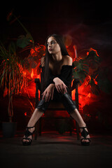 Beautiful girl in dark wear posing on the old retro style chair on a green plants background.