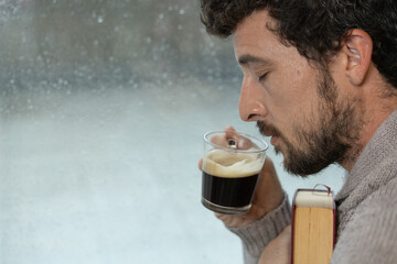 Young adult man enjoys hugging a book and having a cup of warm coffee in his living room at home next to the window on a rainy day, warm at home in autumn or winter, enjoying reading, reading book