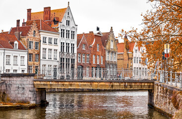 Naklejka premium Bruges, Belgium. Medieval vintage stone bridge and antique brick houses, covered red leaves of trees. Fall autumn landscape. In Brugge.