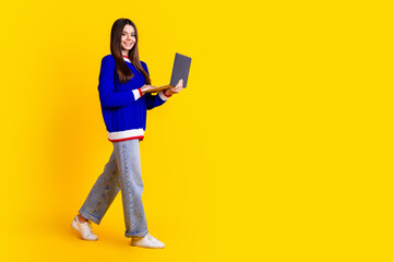 Full length photo of pretty teen girl hold netbook walking wear trendy knitwear blue outfit isolated on yellow color background