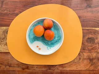 Top view of three fresh peaches in a ceramic blue bowl, placed on an orange placemat over a wooden table, creating a vibrant and colorful contrast.