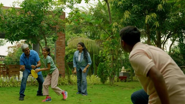 Indian Asian family of four spending quality time outdoors in their home lawn, parents in their forties watching as teenage son teaches his little sister how to throw a frisbee or flying disc