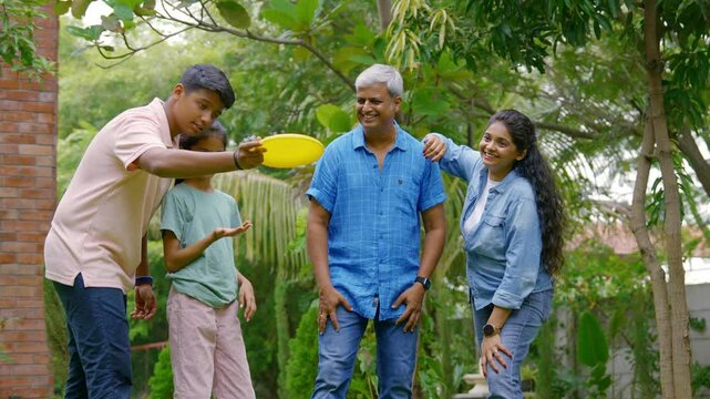 Indian Asian family of four spending quality time outdoors in their home lawn, parents in their forties watching as teenage son teaches his little sister how to throw a frisbee or flying disc