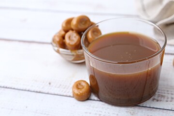 Tasty caramel sauce in bowl and candies on white wooden table, closeup. Space for text