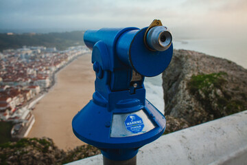 Fototapeta premium A blue public telescope overlooks a beach and coastal town below, offering a panoramic view of the shoreline and rooftops under a cloudy sky