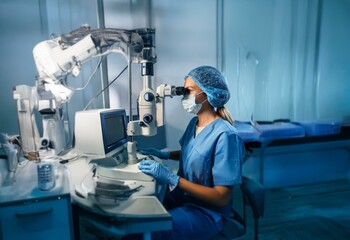 A female surgeon in blue scrubs and a surgical cap uses a surgical microscope in an operating room.