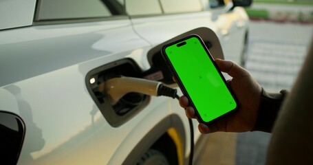 Fototapeta premium A man holds a mobile phone with green screen in his hand, standing near an electric car. Close-up of his hands. Charging cable in the connector