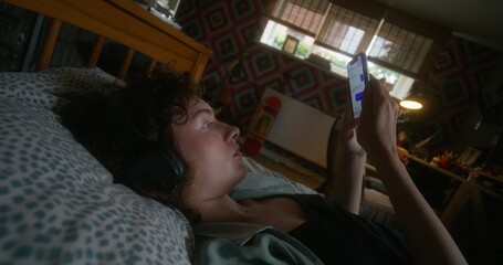 A young woman with headphones on her head sends text messages on a mobile phone lying on a bed with a depressed look.