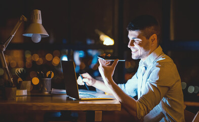 Young Entrepreneur Using Voice Assistant On Phone Working On Laptop Computer In Night Office. Business Lifestyle Concept