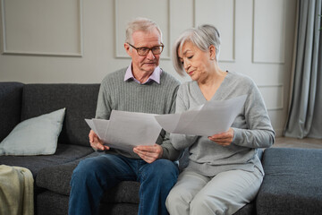 Sad tired disappointed middle aged senior couple sit with paper document. Unhappy older mature man woman reading paper bill managing bank finances calculating taxes planning loan debt pension payment