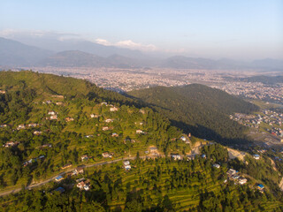 A scenic aerial view of a mountainous village surrounded by lush greenery and distant cityscape.