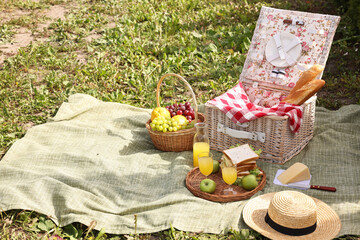 Picnic basket with tableware, different snacks, juice and hat outdoors, space for text