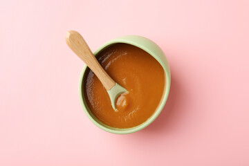 Delicious baby food with spoon in bowl on pink table, top view