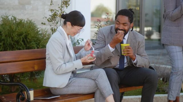 Black businessman and Asian businesswoman doing lunch break outdoors from office building sitting on a bench