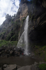 A person standing at the base of a towering waterfall surrounded by rocky terrain and greenery.