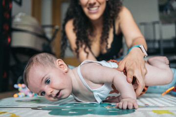 Mother helping baby girl lying on tummy lifting herself. Mother smiles watching her baby girl strengthen her neck and back muscles by lifting her upper body