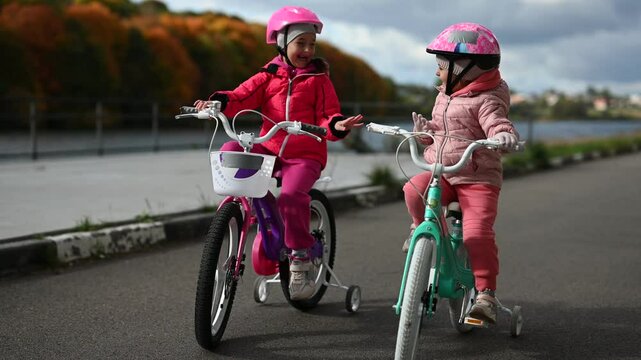 Two happy little girls are riding their bicycles on a paved path beside a river. They're wearing helmets and having fun together on a beautiful autumn day.