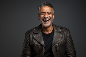 Obraz premium Portrait of a cheerful indian man in his 40s sporting a classic leather jacket while standing against soft gray background